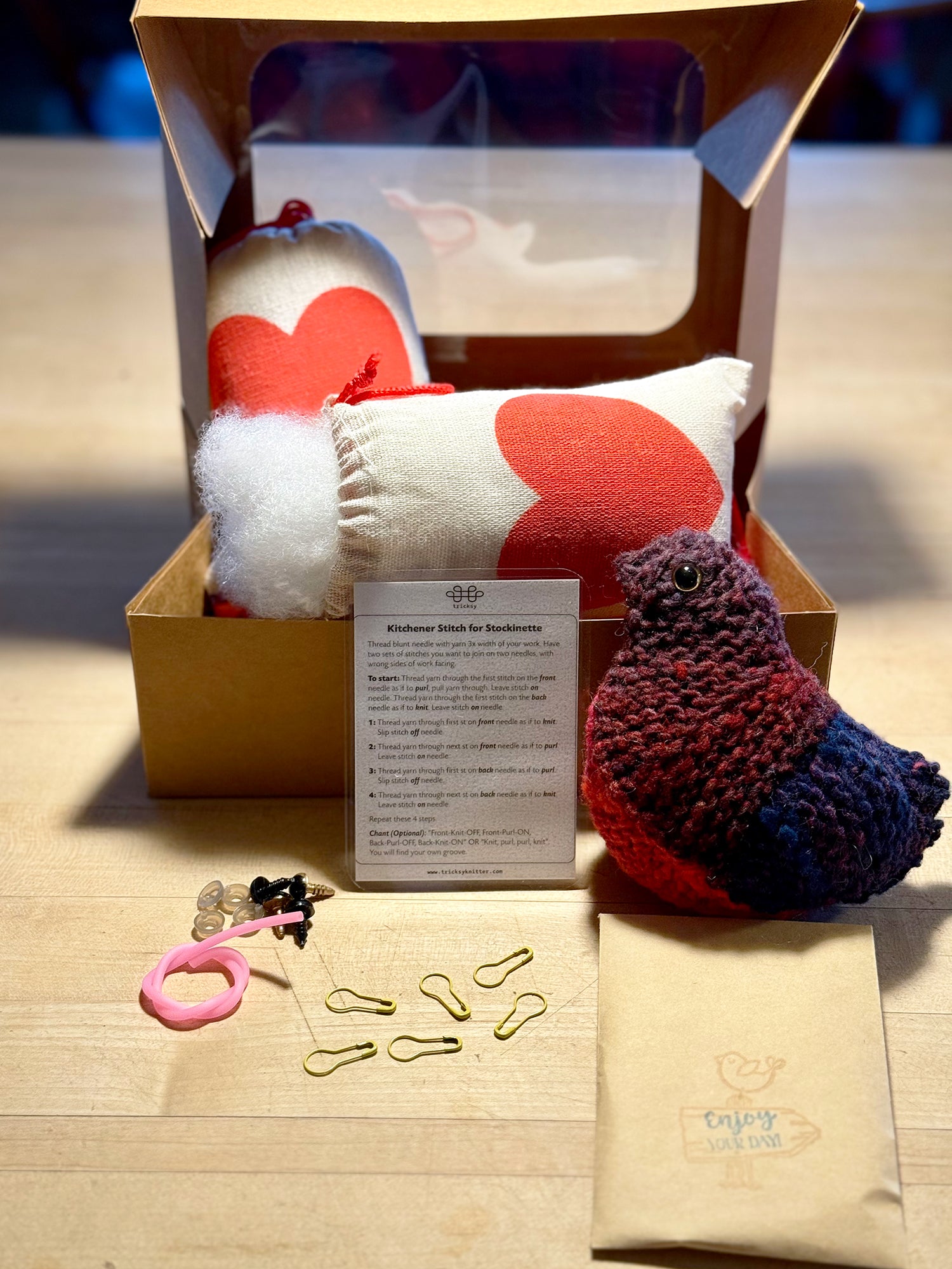 Knitted items including a heart-shaped pillow, a bird, and a package on a wooden surface.